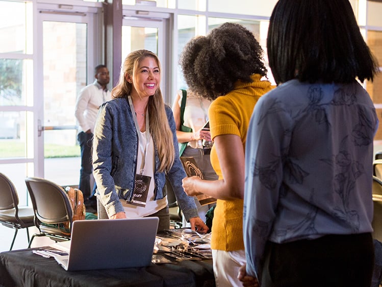 People gathered around a registration table in a bright event venue. Attendees talk and move through the space with rows of chairs and large windows in the background.