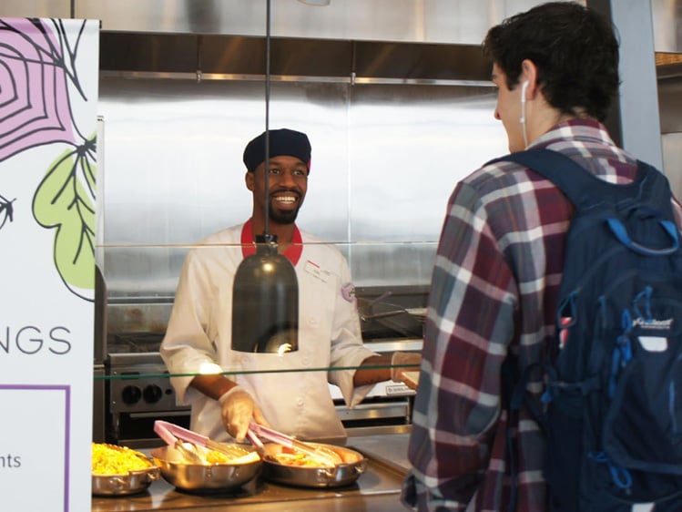 Person with a backpack standing at a cafeteria serving station, interacting with a food service worker behind a counter with hot food trays, beside signage displaying ingredient and allergen information.