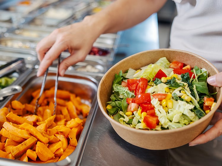 Person assembling a fresh salad bowl from a self-serve bar with vegetables and toppings.