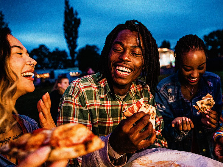 Group of people gathered outdoors in the evening, sharing pizza and food while standing together in a casual social setting with a grassy area and buildings in the background.