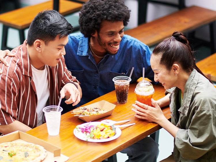 Three people seated at a cafeteria-style table sharing pizza, salads, and drinks, gathered around a wooden bench table in a bright casual dining space.