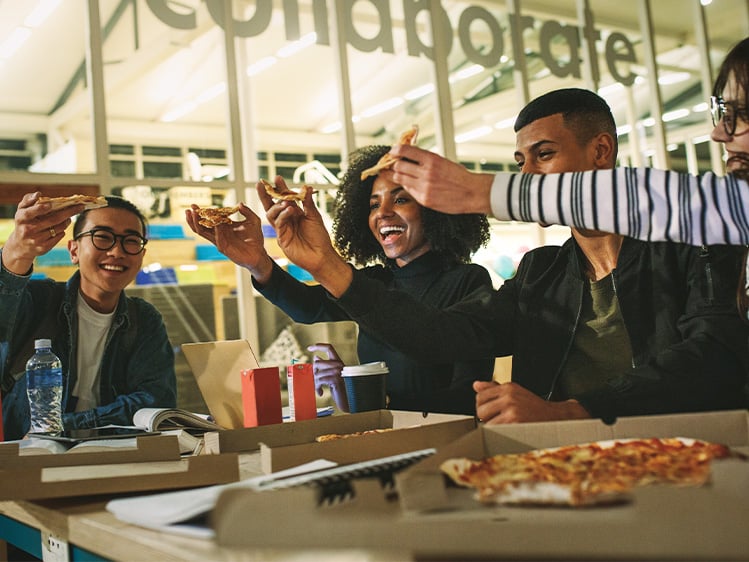 Group of students gathered around a table sharing pizza slices and drinks, with study materials laptops and food boxes visible inside a brightly lit campus dining area.