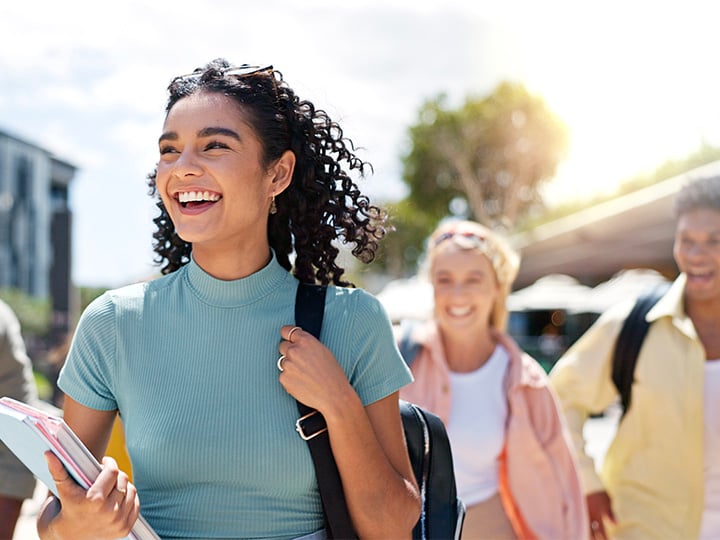 Young person walking outdoors on a sunny day, carrying notebooks and a backpack, with other students walking behind across a campus or urban walkway.