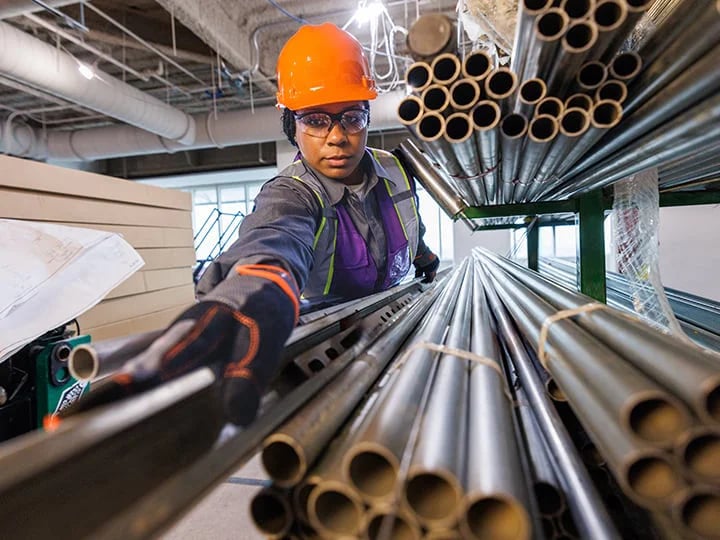 Construction worker in orange hard hat and gloves reaches for a metal pipe from a stack, surrounded by building materials, ceiling ducts, and lighting fixtures.