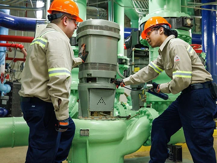 Two workers in safety helmets and uniforms perform maintenance on industrial machinery with green pipes and components, using tools to adjust equipment.