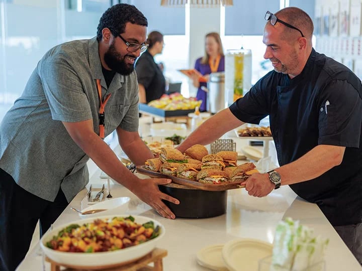 Two people arrange sandwiches on a buffet table with assorted dishes at a catered event.
