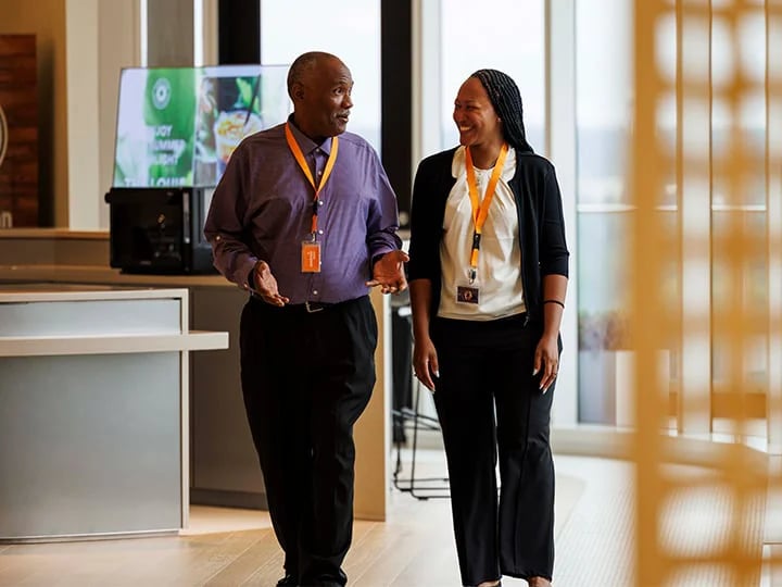 Two people with lanyards walk and talk in a modern office with large windows and a digital screen.