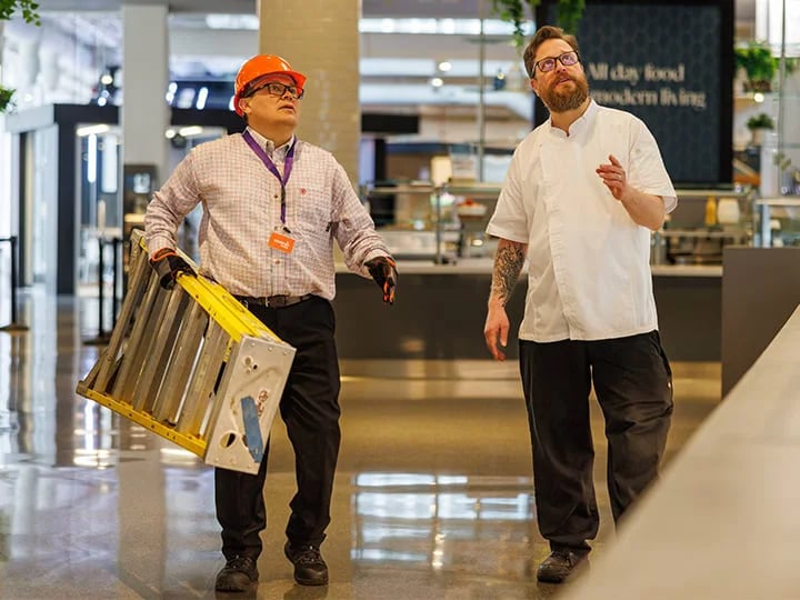 Two individuals converse in an indoor commercial space; one holds a step ladder wearing safety gear, the other in a chef’s coat.