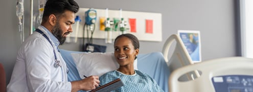 A hospital worker checking a chart next to a patient sitting in bed smiling