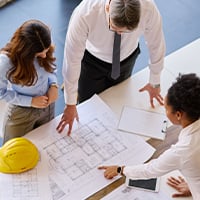 Team reviewing architectural blueprints on a table, pointing at plans during a discussion, with a construction hard hat and documents visible in a collaborative workspace.