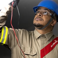 Technician wearing a hard hat, safety glasses, and protective gear inspecting electrical wiring with test leads against a dark background with visible cables.