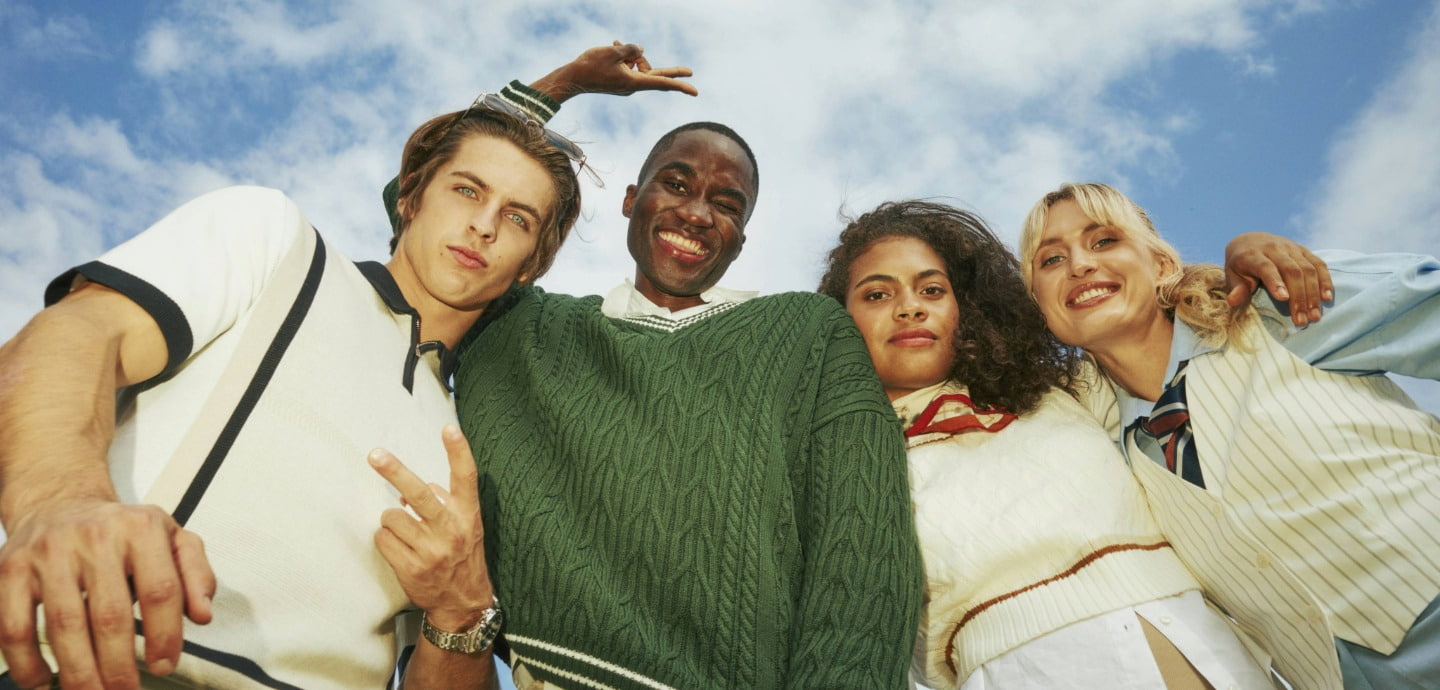 Four friends smiling and leaning toward the camera from a low angle under a blue sky.