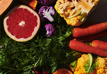 Assorted fresh produce including halved grapefruit, purple and yellow cauliflower, carrots with greens, and leafy greens arranged in a colorful display.