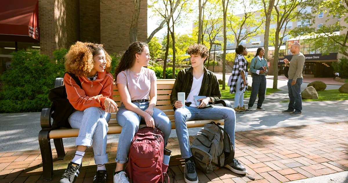 College students sitting on a bench outdoors on campus, with other students standing in the background.