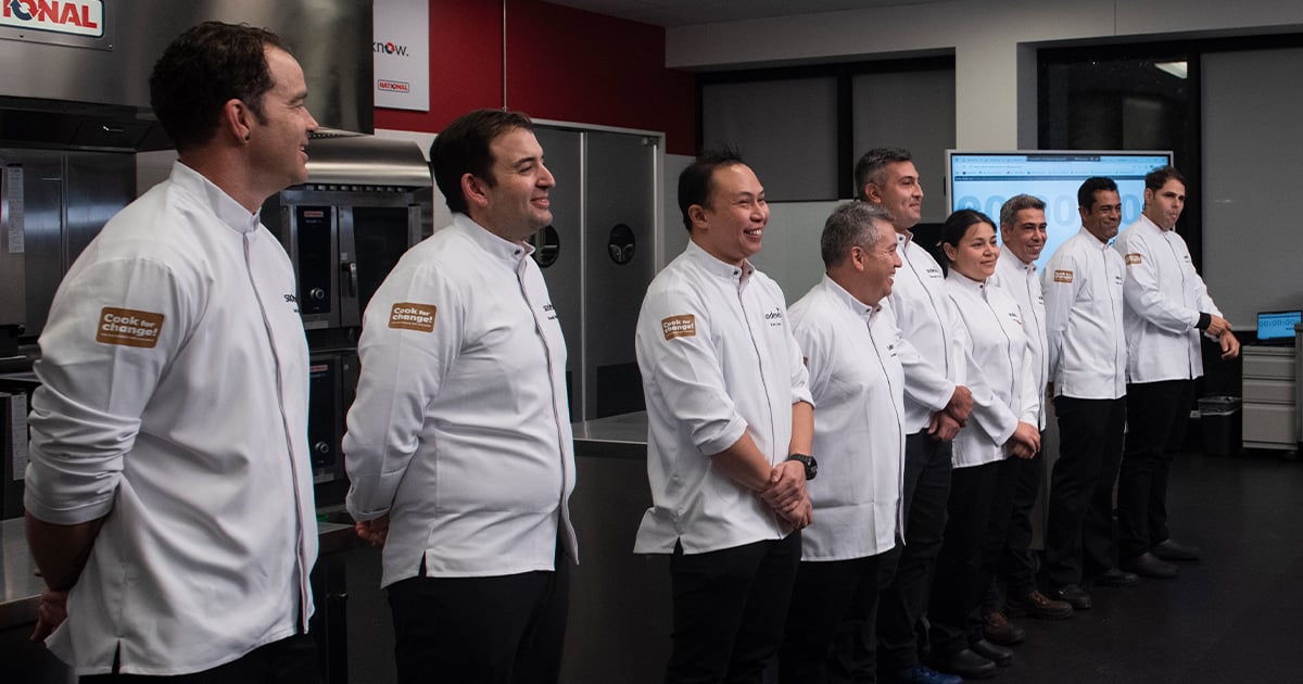 Seven chefs in white jackets with 'Cook for Change' patches standing in a commercial kitchen near equipment.