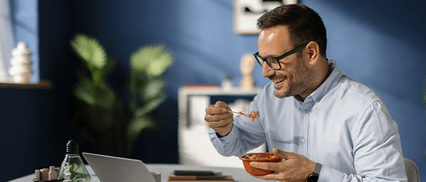 Person eating breakfast at a desk while working on a laptop in a bright, modern home office.