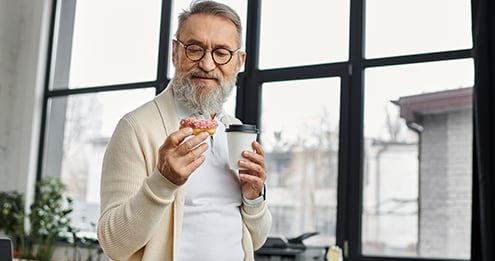Person holding a coffee cup and a pink-frosted donut while standing near large windows in a bright indoor space.