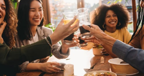 People clinking glasses of dark beverages around a wooden table with bowls of food in a bright indoor setting.