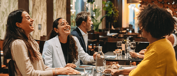 Group of people sitting at a restaurant table, talking and enjoying drinks in a warm, modern dining setting.