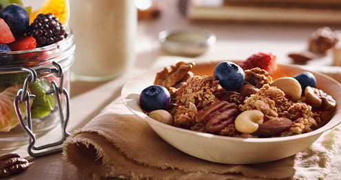 Bowl of granola with nuts and blueberries next to a jar of mixed fruit and a glass of milk on a rustic cloth.