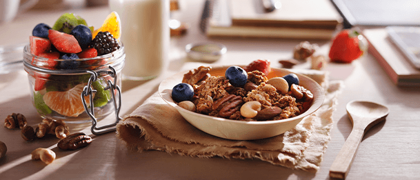 Bowl of granola with berries beside a jar of mixed fruit and a glass of milk on a breakfast table.