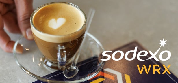 Hand holding glass cup of espresso with heart latte art on saucer beside Sodexo WRX logo and booklet.