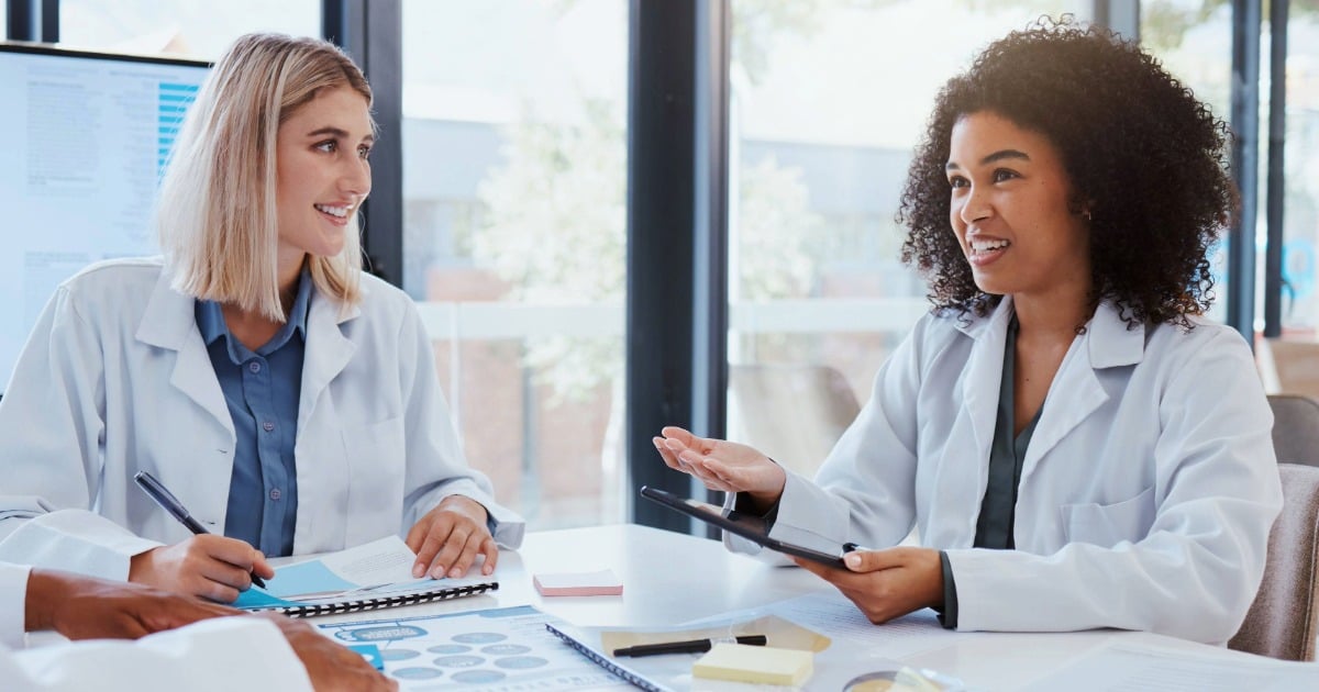 Two dietitians sit at a table reviewing research notes, with one holding a tablet and data displayed on a screen behind them.