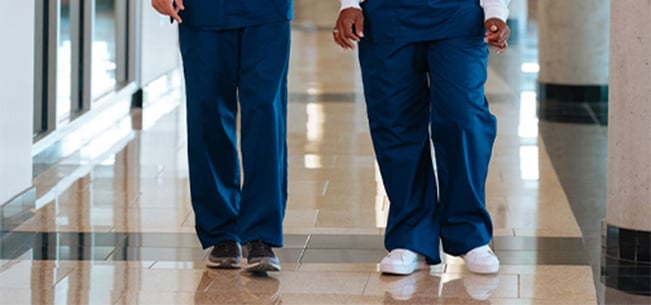 Two healthcare workers walking down a polished hospital corridor wearing blue medical scrubs.