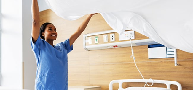 Healthcare worker making a hospital bed by lifting a clean sheet in a patient room.
