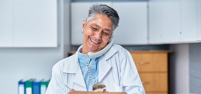 Healthcare professional on a phone call while writing notes on a clipboard in a medical office.