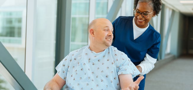 Patient in a wheelchair with a healthcare worker walking alongside in a hospital corridor.