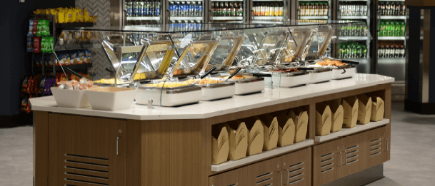 Buffet-style food station with hot dishes under glass and shelves stocked with takeout containers.