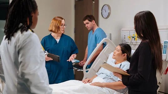 An interdisciplinary care team standing around a patient’s hospital bed, discussing the patient’s care plan.