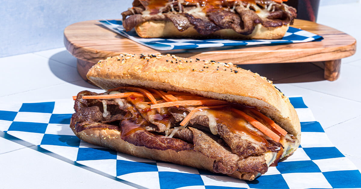 Close-up of two toasted sandwiches filled with sliced beef, shredded vegetables, and sauce, placed on blue checkered paper with a wooden board in the background.