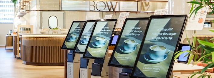 Row of touchscreen ordering kiosks displaying coffee menus inside a bright, modern café interior.