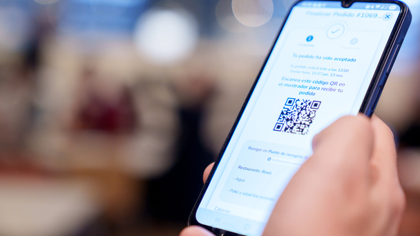 Smartphone displaying a scan-and-go order confirmation with a QR code, held in a café setting.