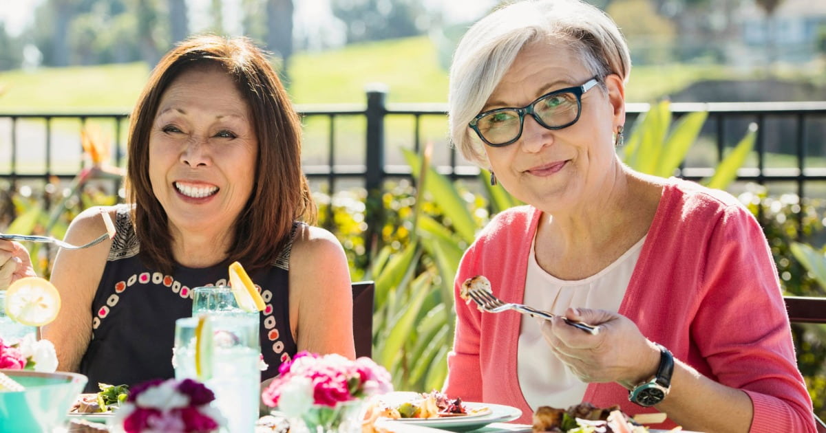 Two older adults sit outdoors at a sunlit table enjoying a meal together, surrounded by greenery and flowers.