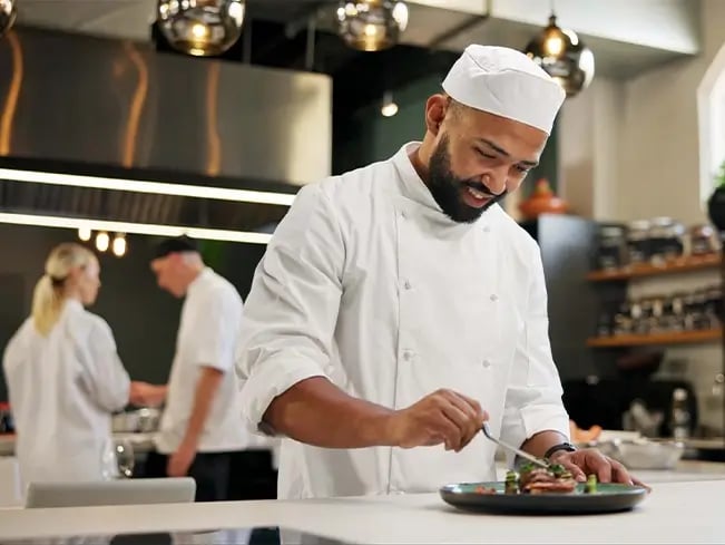 Three chefs in white uniforms work in a modern kitchen with stainless steel appliances; one chef in the foreground plates food while others work in the background