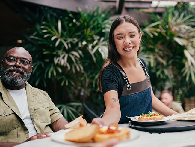 Two people sit at an outdoor table with greenery in the background; one in a black shirt and apron serves a plate with toast and salad or sandwich to the other.