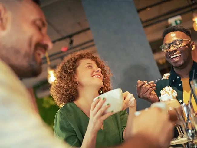Group seated at a café table; one person with curly hair in a green top holds a white cup, another with short hair and glasses eats dessert with whipped cream; café interior in background.