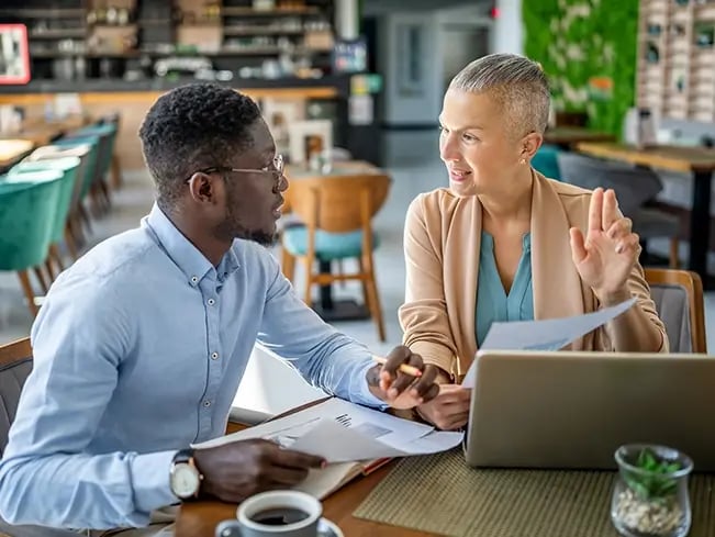 Two people sit at a table in a cafe or restaurant, discussing documents; one gestures while the other holds papers. A laptop, coffee cup, and papers are on the table.