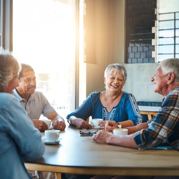 A group of seniors sitting around a café table, smiling and chatting as they each enjoy a cup of coffee.