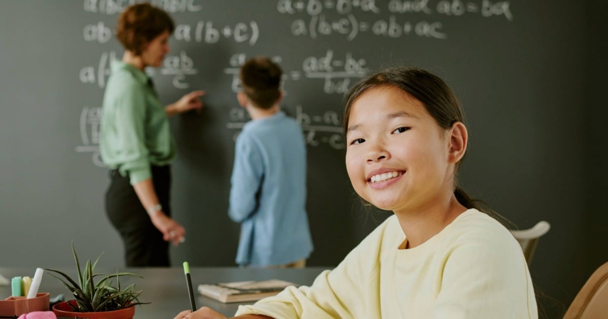 A student sitting at a desk in a classroom while a teacher and another student work on math equations at the chalkboard in the background.