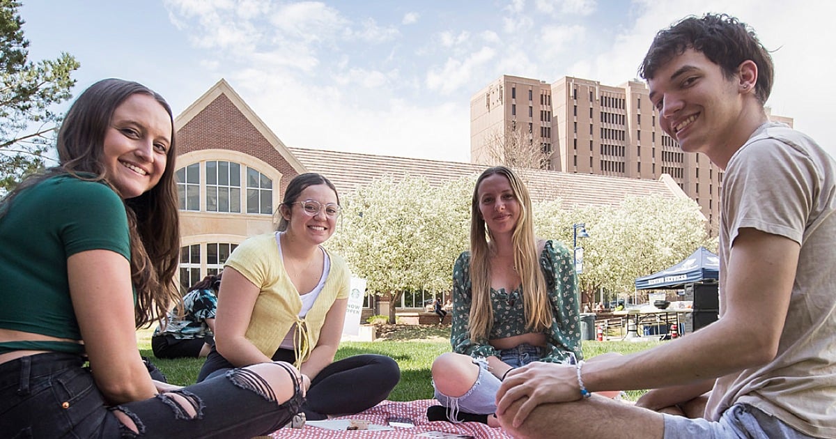 A group of UNC students sitting together outdoors on campus, smiling at the camera as they eat lunch.