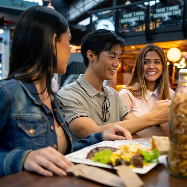Three people dining together at a food hall table, sharing a meal and conversation.