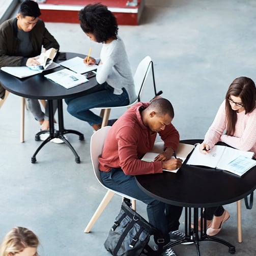 Students seated at tables, studying, writing notes, and reviewing coursework in a shared learning space.