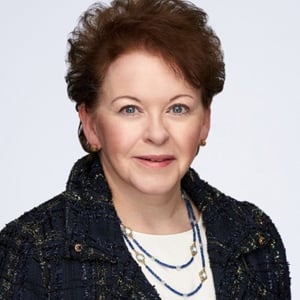 Studio headshot of a person with short dark hair, wearing a navy textured jacket and layered necklaces on a light background