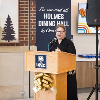 Person speaking at a UNC podium during a Holmes Dining Hall event, with signage and ribbon decoration.
