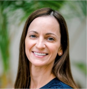 Person with long brown hair wearing a dark top, standing in front of green foliage.