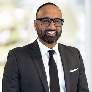 Head-and-shoulders portrait of a person in glasses wearing a dark suit and tie against a light, blurred background.